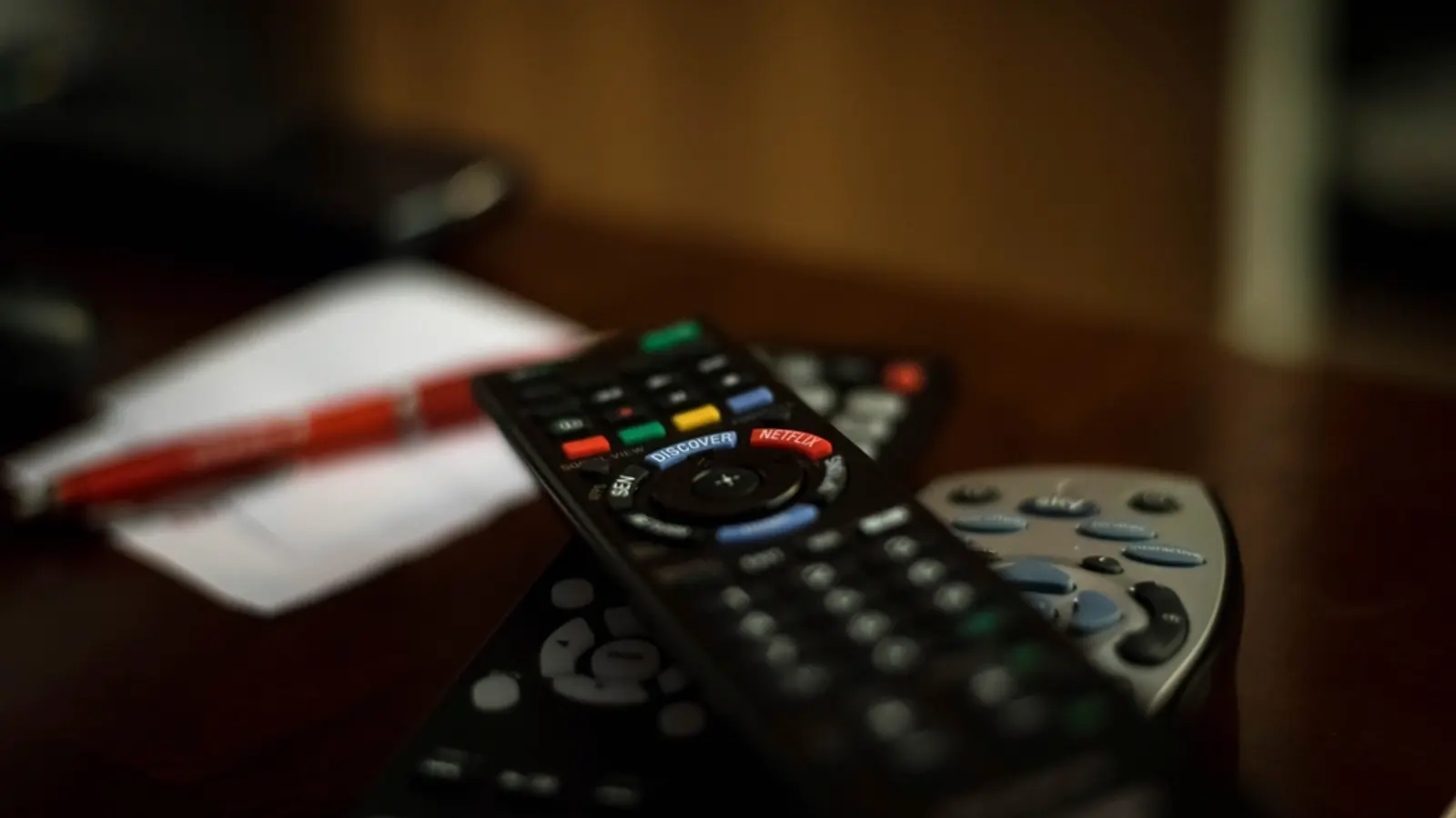 Two TV remotes lying on a dark table beside a notepad