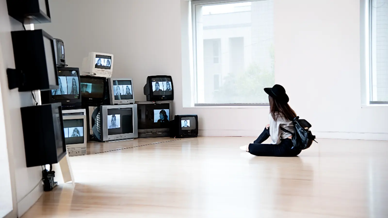 A woman sitting on the floor of a bright room surrounded by old TV sets