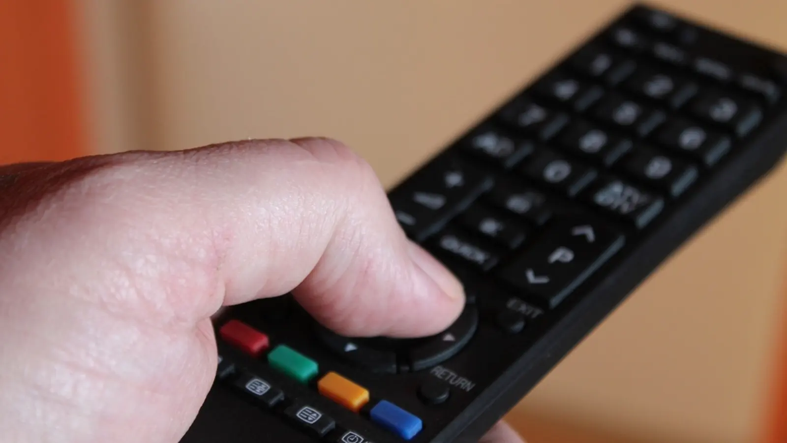 A hand holding a black TV remote with coloured buttons