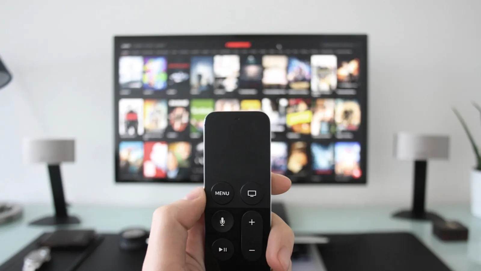 Hand holding a streaming remote in front of a TV showing a grid of shows